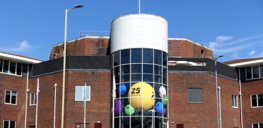 Allwyn, dříve Sazka, loni v ČR zvýšil zisk na 2,1 mld. Kč a tržby na 14,3 mld. Brick office building with a cylindrical glass atrium filled with oversized colorful lottery balls visible through the windows ch:.25 central yellow ball