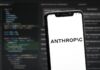 Šéf podniku Anthropic vyzval firmy, aby zpomalily vývoj modelů umělé inteligence Smartphone angled on a blurred code editor background showing the word ANTHROPIC on a white screen.