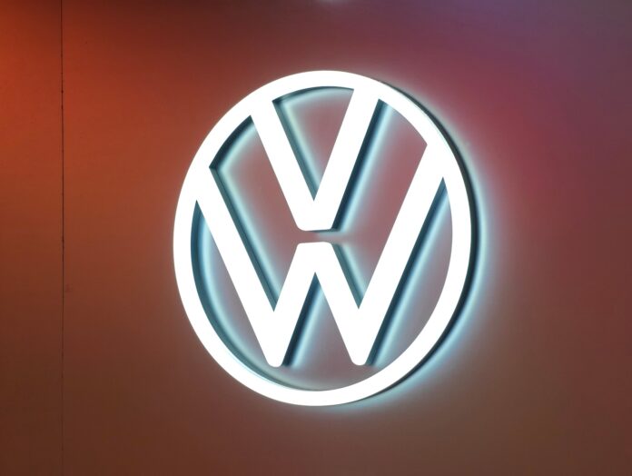 Illuminated Volkswagen logo (white VW circle) on a warm orange wall background.