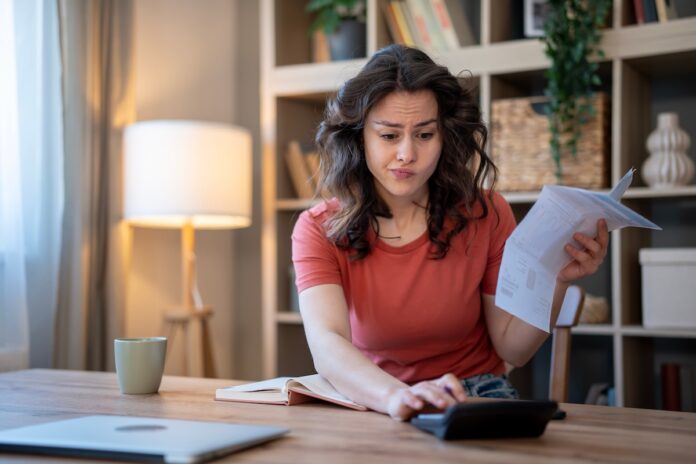 Woman at a wooden table, holding papers and using a calculator, looking puzzled.
